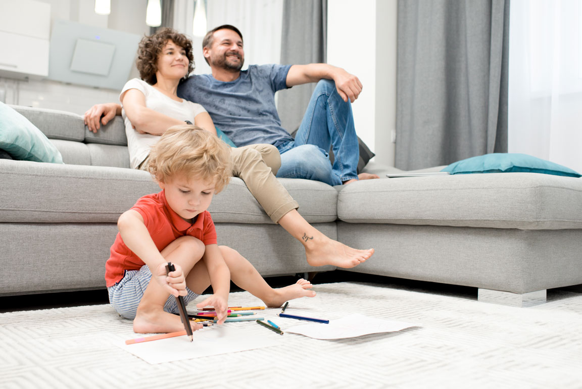 husband and wife sitting on living room couch with their son drawing on the floor