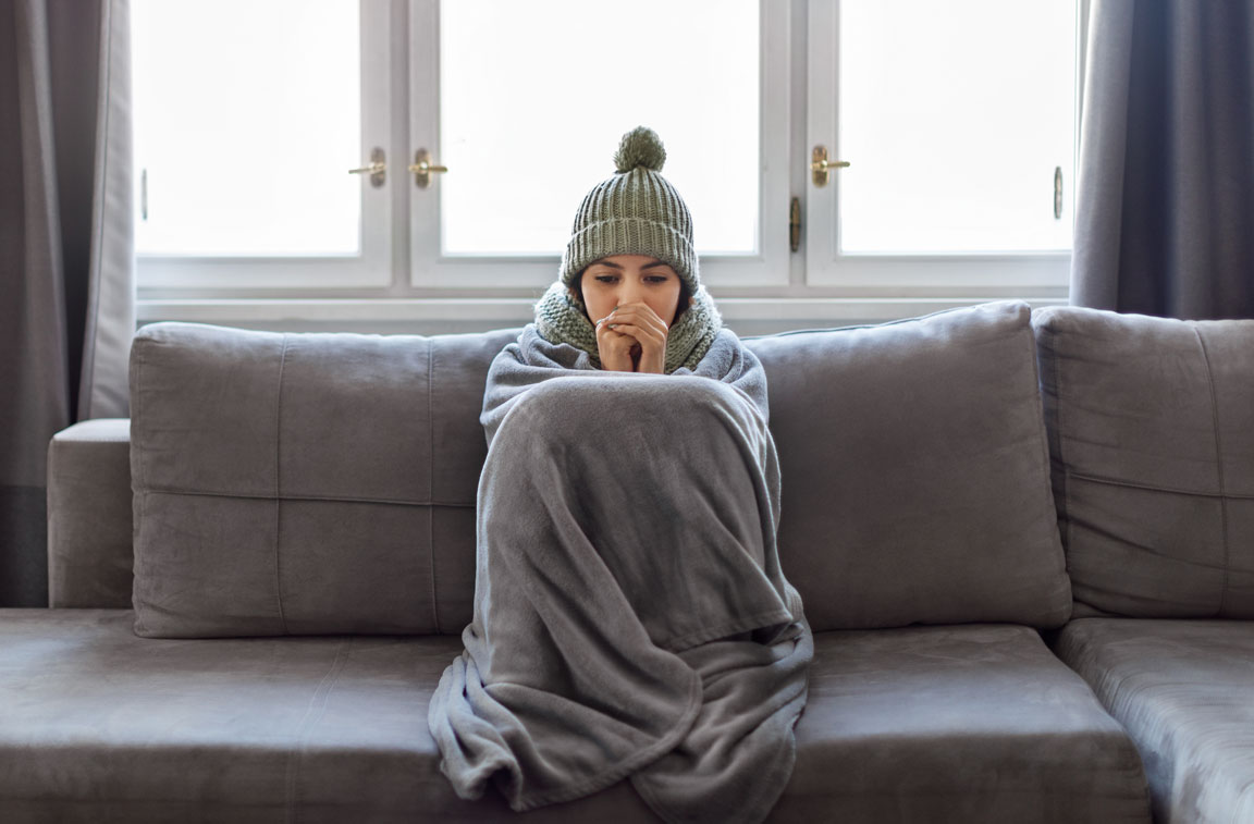 cold woman sitting on couch bundled up in blanket and winter hat